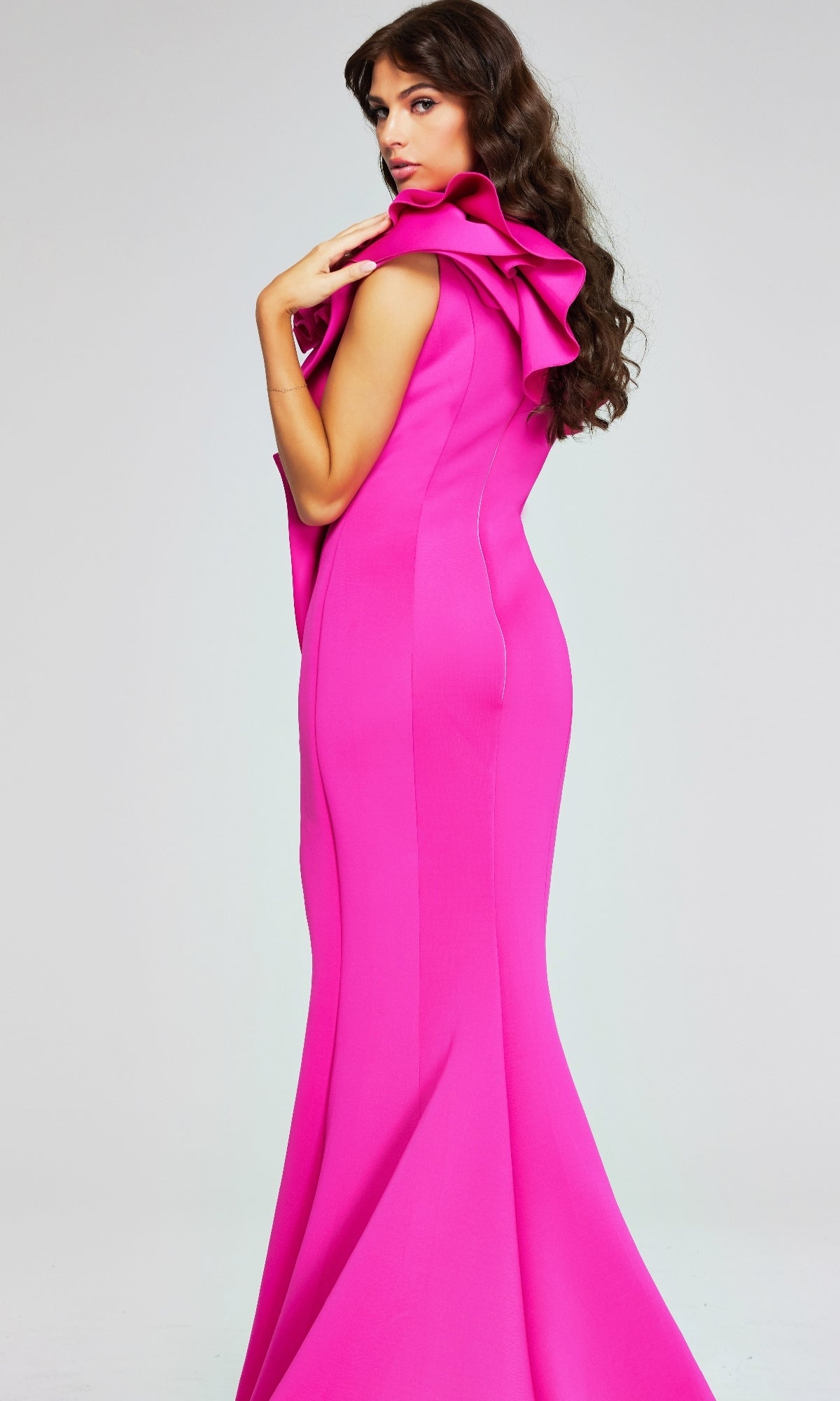 A woman with long wavy hair models the Jovani 37400, a bright pink, form-fitting long formal dress with a dramatic ruffled one-shoulder neckline, posed against a plain light background.